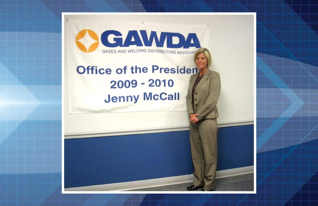 Professional woman in a beige pantsuit stands beside a banner reading 'Office of the President 2009–2010 Jenny McCall' at a GAVDA event/aligned with the banner.",