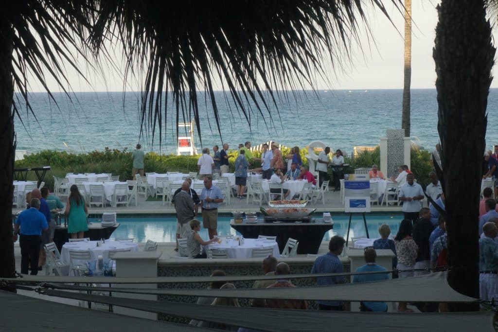 Seaside outdoor party with guests around white-clothed tables and buffet, ocean view in the background.