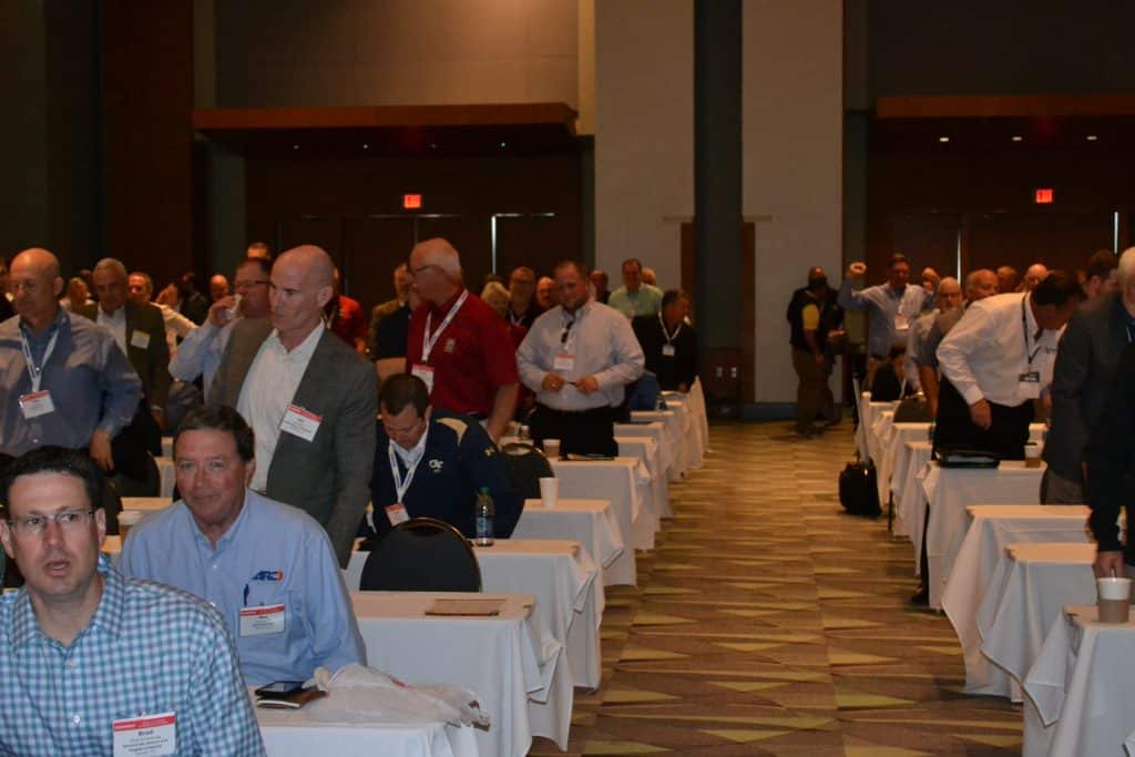 Crowded conference room with attendees wearing name badges standing and talking around rows of white-draped tables.