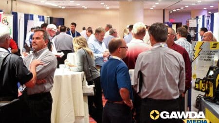 Crowded trade show floor with attendees chatting near booths and banners, badges visible.