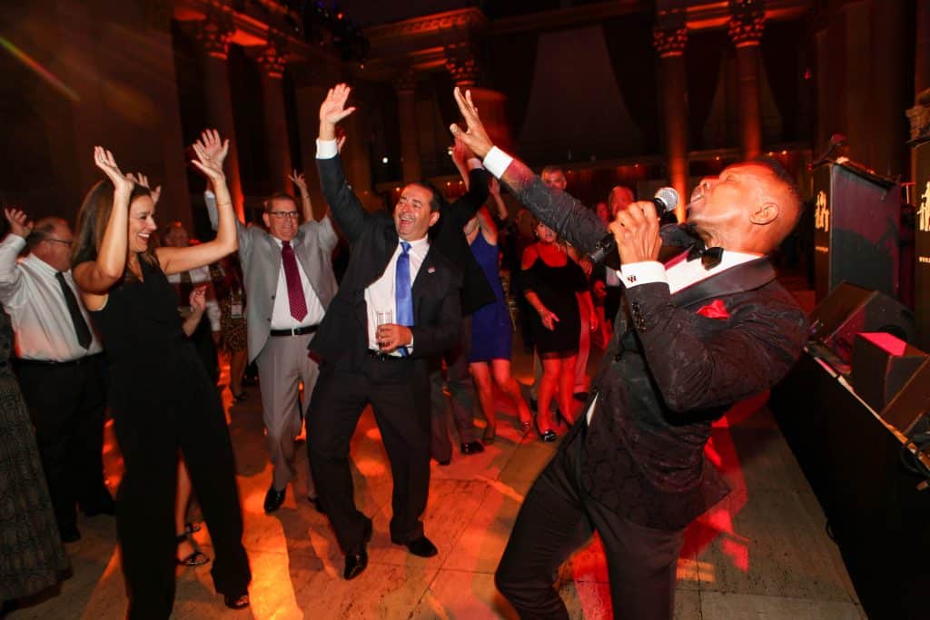 People in formal attire dancing and cheering at a party under warm red lighting, with a singer on the right performing into a microphone.