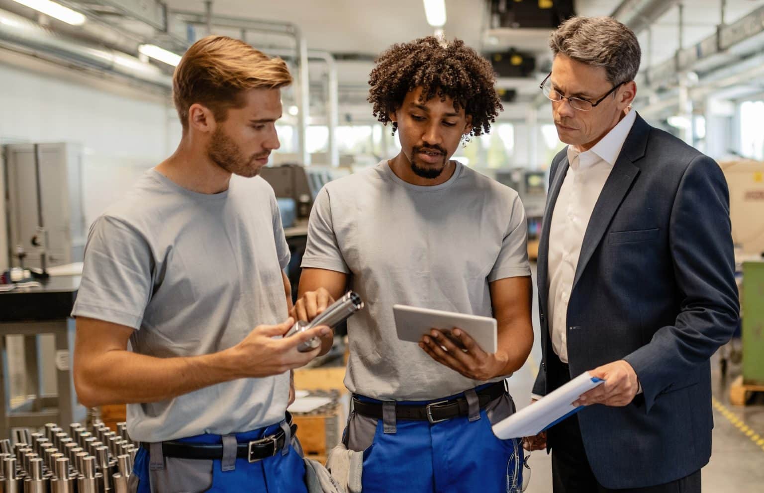 Three colleagues in a factory discuss a project: two workers in grey shirts and blue pants examine a metal part and tablet while a suited supervisor reviews blueprints.