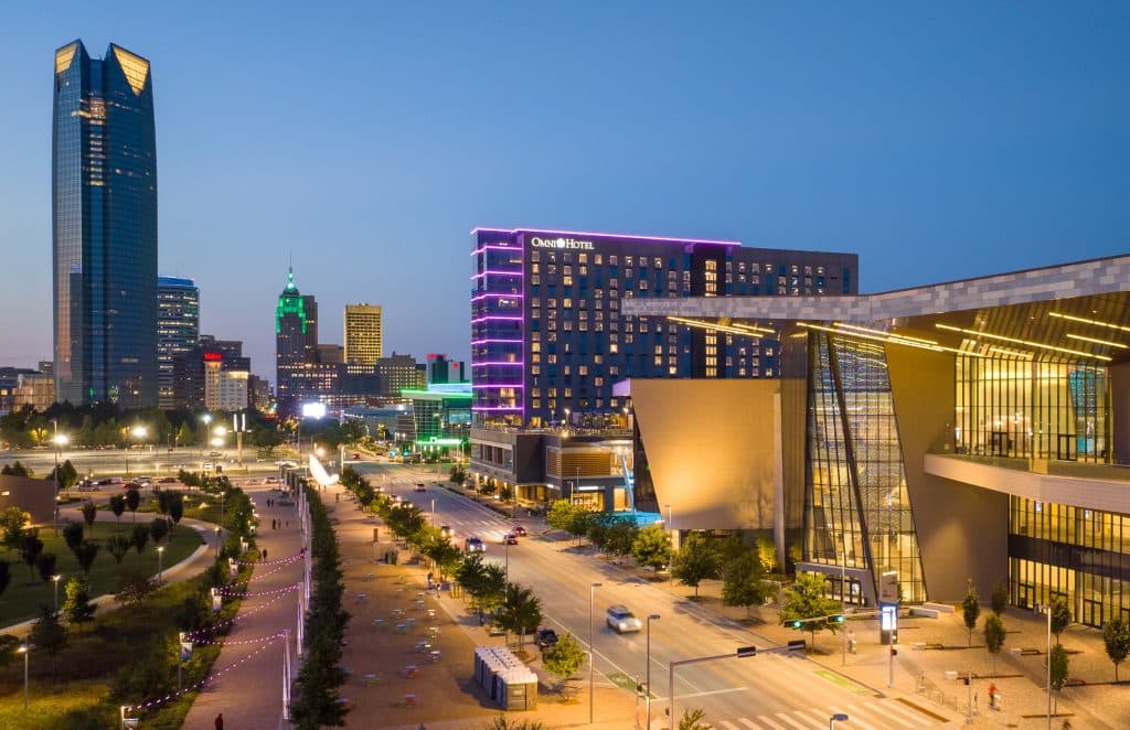 Dusk city skyline with a tall glass skyscraper left, a neon-lit Omni Hotel in the center, and a modern structure in the foreground.