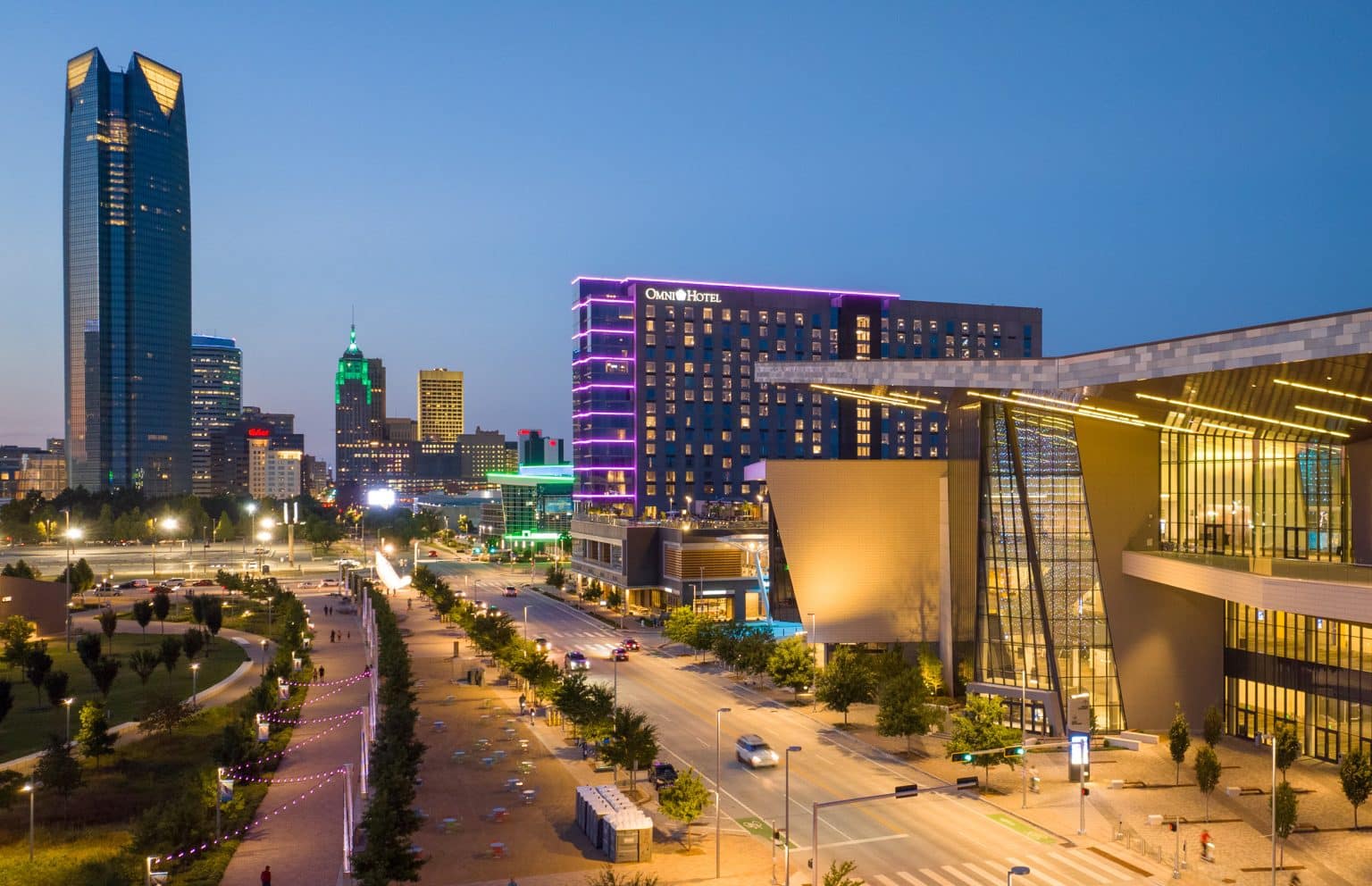 Dusk city skyline with a tall glass skyscraper left, a neon-lit Omni Hotel in the center, and a modern structure in the foreground.