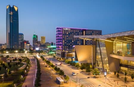 Dusk city skyline with a tall glass skyscraper left, a neon-lit Omni Hotel in the center, and a modern structure in the foreground.