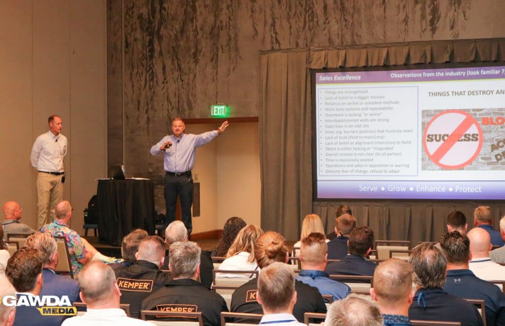 Two men present on a stage in a conference room; one speaks into a microphone while gesturing to a large slide titled 'Sales Excellence'.