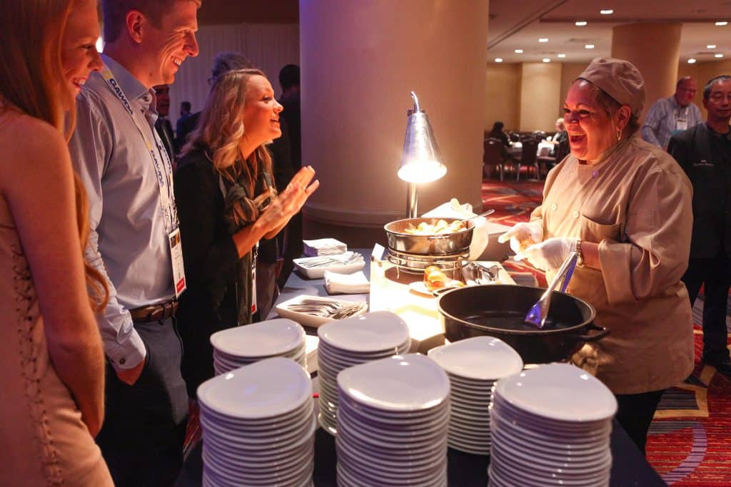 Chef in beige uniform serving food at a buffet as guests wait with plates nearby.