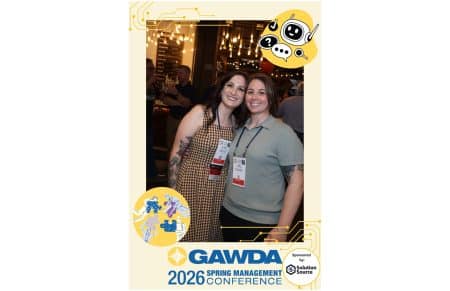 Two women wearing conference badges smile for a photo at a busy event with decorative graphics in a venue backdrop.