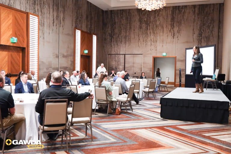 Speaker on a stage addresses professionals seated at round tables in a banquet-style conference room.