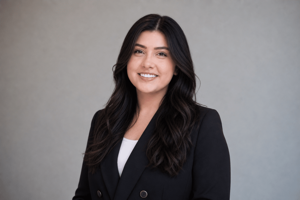 Professional headshot of a smiling woman with long dark hair in a black blazer against a gray backdrop.