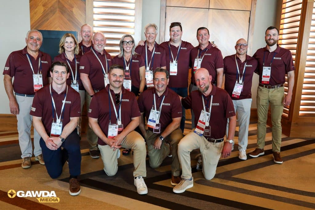 Group of thirteen people in maroon polo shirts with name badges posing for a photo in a lobby; GAWDA MEDIA logo visible in the bottom left.