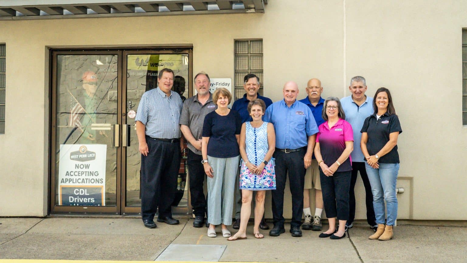 Group of eleven adults posing outside a beige building with a glass door entrance. Signs on the door advertise CDL driver jobs and accepting applications.