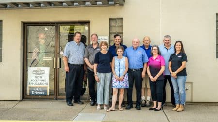 Group of eleven adults posing outside a beige building with a glass door entrance. Signs on the door advertise CDL driver jobs and accepting applications.