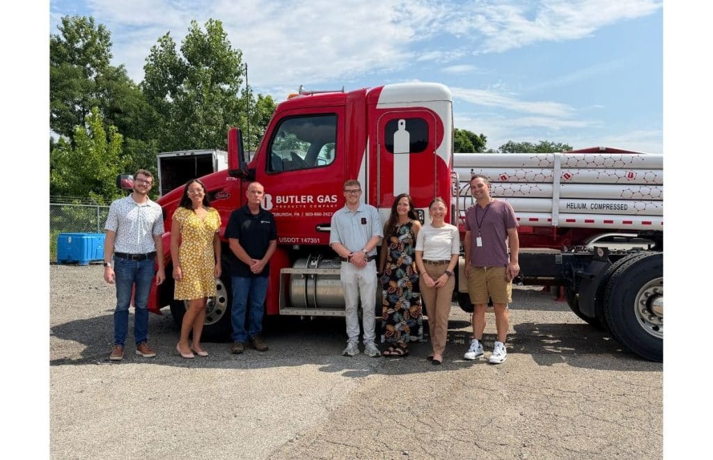 Seven adults stand in a row outdoors beside a red Butler Gas tanker truck with a helium tank in the background.