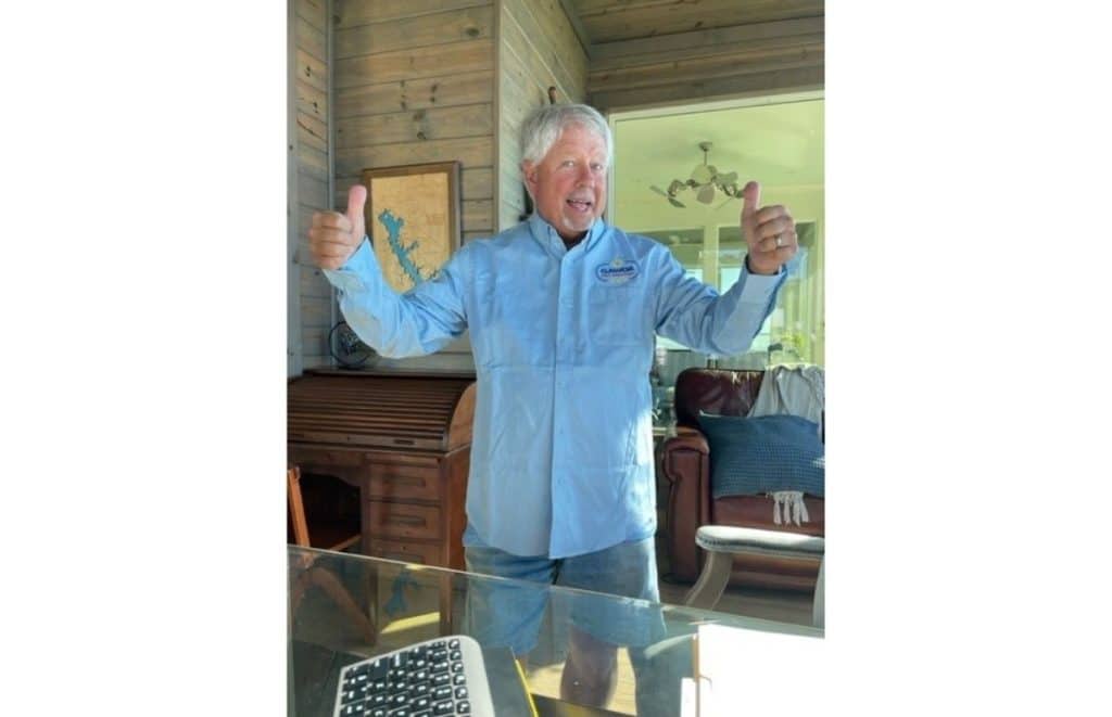 Man in a light blue shirt gives two thumbs up indoors on a wooden porch, smiling at the camera.
