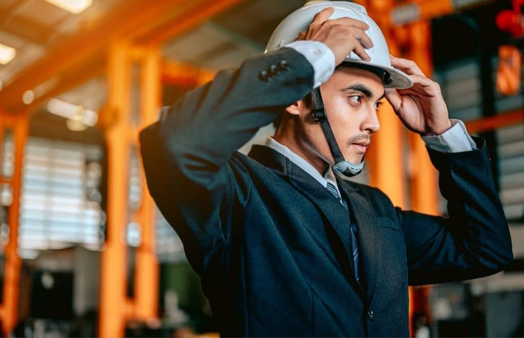 Man in a suit adjusts a hard hat in an industrial warehouse setting.