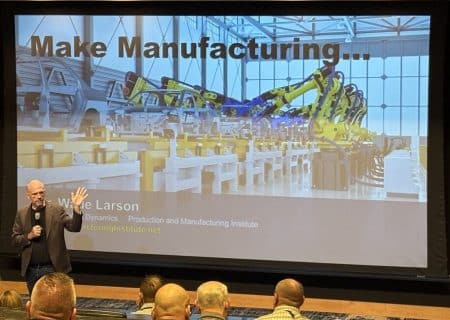 Presenter on a stage delivering a talk; a large screen behind shows a factory with yellow robotic arms and the heading 'Make Manufacturing...', plus the name 'Wade Larson' at the bottom.