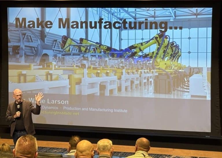 Presenter on a stage delivering a talk; a large screen behind shows a factory with yellow robotic arms and the heading 'Make Manufacturing...', plus the name 'Wade Larson' at the bottom.