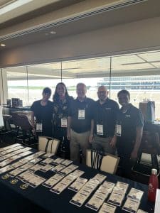 Five people with lanyards stand behind a table covered in rows of name badges, at a vendor booth inside a stadium concourse.