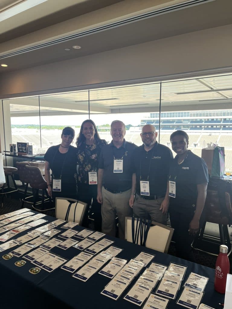 Five people with lanyards stand behind a table covered in rows of name badges, at a vendor booth inside a stadium concourse.