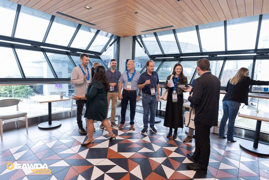 Group of event attendees with lanyards converse near round tables in a glass-walled atrium with geometric tile floor.