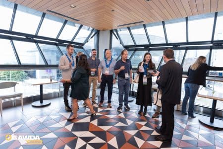 Group of event attendees with lanyards converse near round tables in a glass-walled atrium with geometric tile floor.