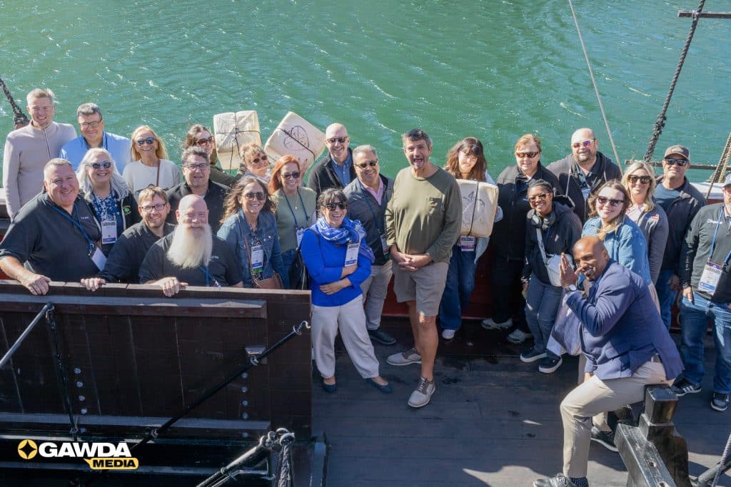 Group of about 25 people on a wooden boat deck posing for a group photo with turquoise water behind them; many wear lanyards and casual outdoor clothing. GAWDA Media logo visible bottom left.