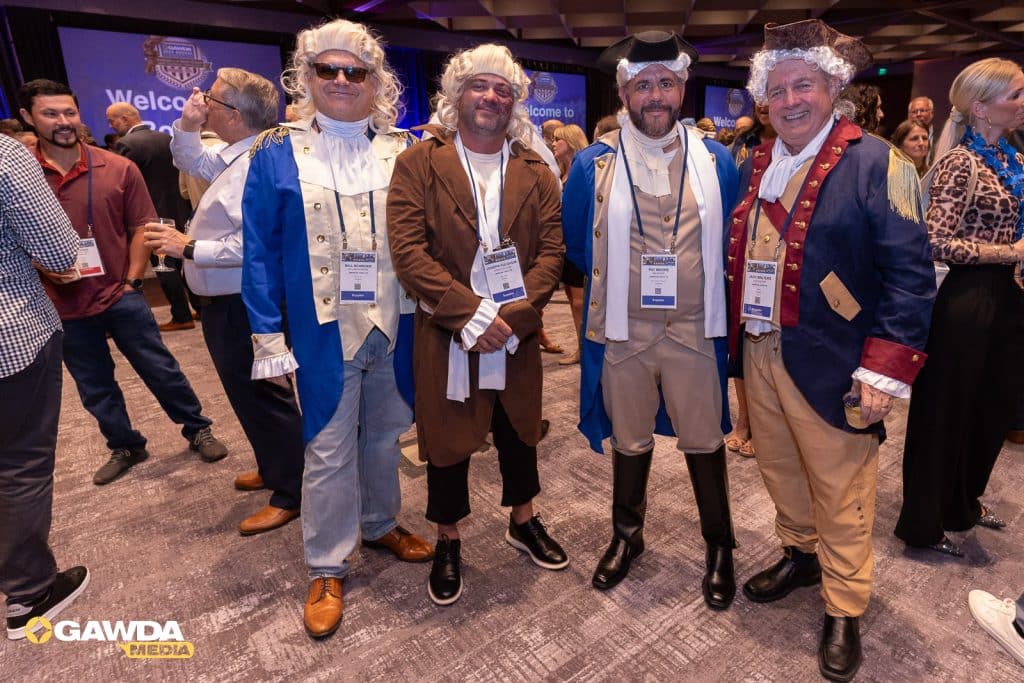 Four men in 18th‑century costumes pose for a photo at a conference, wearing wigs, coats, and name badges; a crowd and GAWDA logo are in the background.
