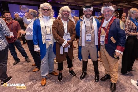 Four men in 18th‑century costumes pose for a photo at a conference, wearing wigs, coats, and name badges; a crowd and GAWDA logo are in the background.