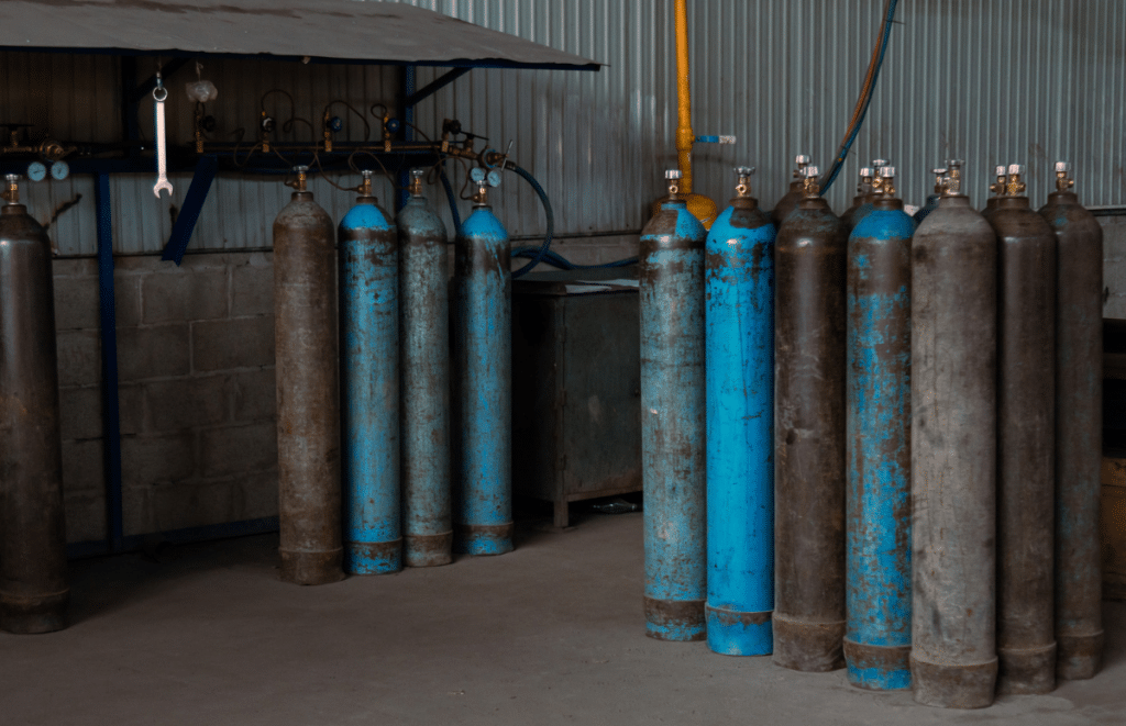 Row of blue and rusted gas cylinders standing upright along a workshop wall with gauges and hoses in the background.