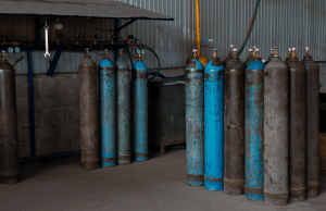 Row of blue and rusted gas cylinders standing upright along a workshop wall with gauges and hoses in the background.
