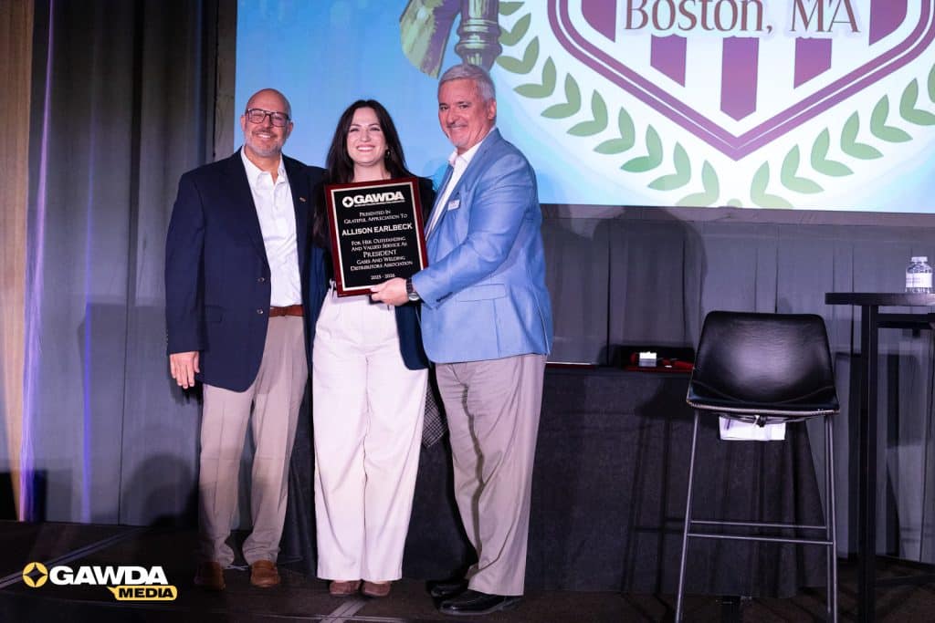 Three professionals on stage pose with an award plaque during a GAWDA event; large emblem backdrop.