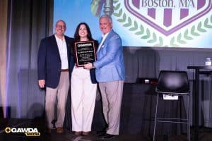 Three professionals on stage pose with an award plaque during a GAWDA event; large emblem backdrop.