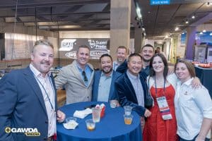 Group of eight smiling professionals posing together at a conference reception around a high-top table with drinks, badges visible, GAWDA Media logo in corner.