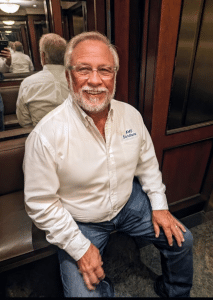 Portrait of a smiling man with gray hair and beard in a white AMS Solutions shirt, seated in a wood-paneled elevator lobby.