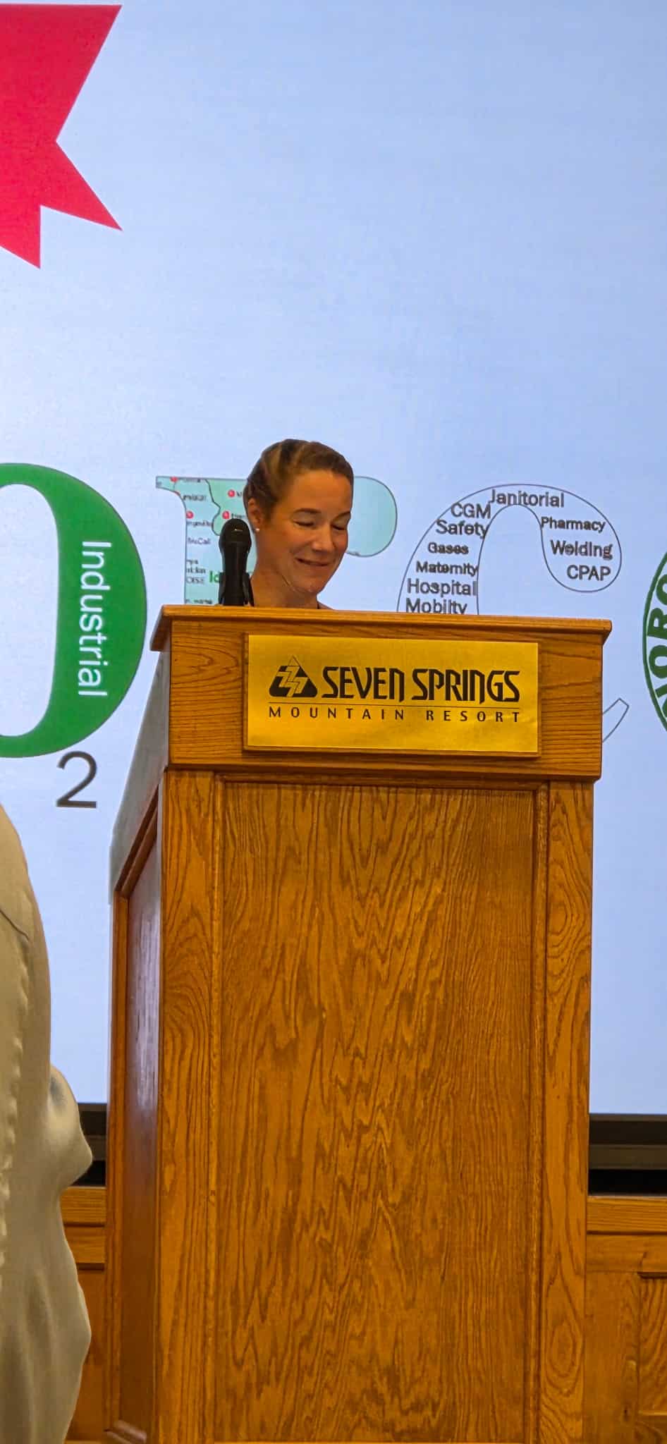 Woman speaking at a wooden podium with a Seven Springs Mountain Resort plaque during a presentation, microphone nearby, with event graphics in the background.