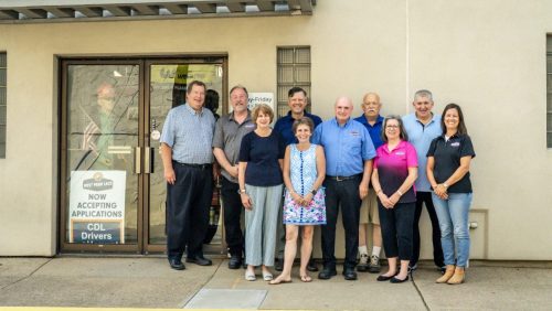 Group of eleven adults posing outside a beige building with a glass door entrance. Signs on the door advertise CDL driver jobs and accepting applications.