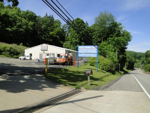 Industrial site with a white metal building, orange truck, and a blue sign advertising Autobody  Welding Materials & Equipment; overhead power lines cross the scene against a clear blue sky with trees in the background.