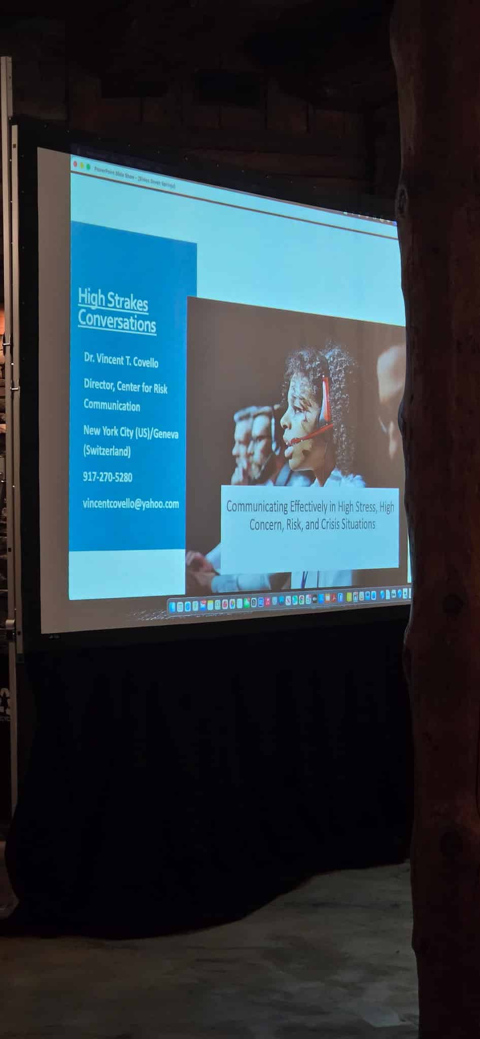 Slide showing 'High Stakes Conversations' with Dr. Vincent Covello's bio and contact info; a speaker wearing a headset appears on the right side in a projected presentation.