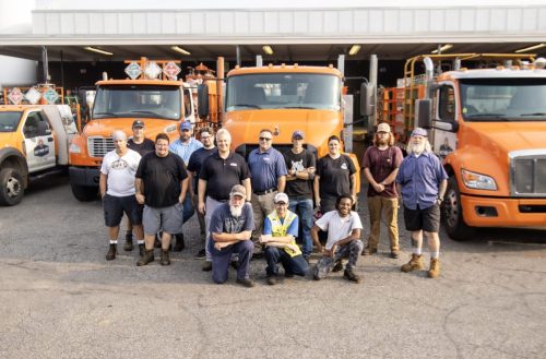 Group of workers posing in front of large orange trucks at a maintenance yard or fleet depot.