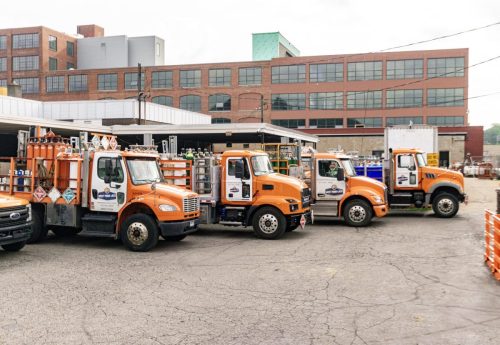 Line of orange utility trucks parked in a loading yard with a brick industrial building in the background, giving an industrial service scene.