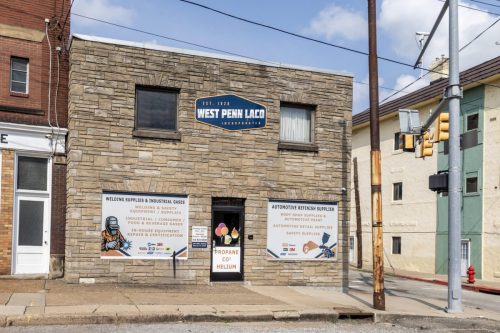 Stone storefront with a blue West Penn Laco sign, two windows, and posters advertising welding and auto supplies on the lower wall.