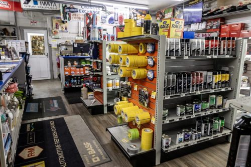 Auto body supply aisle with yellow tape rolls on orange display panels and paint cans on adjacent shelves in a shop.]} }? Wait format expects array of strings. We'll provide multiple. Need proper JSON object. Let's craft: {
