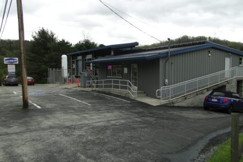 Gray metal building with blue roof and ramped entrance; cloudy sky and trees in the background, with a parking lot in front.