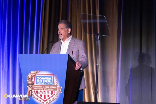 Speaker at a podium delivering a talk at GAWDA 2026 Annual Convention, Boston, MA, with blue and gold stage curtains behind him.