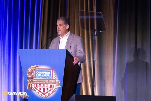 Man in a blazer speaks at a podium on a stage with a GAWDA 2026 Annual Convention banner in Boston, MA, microphone nearby.