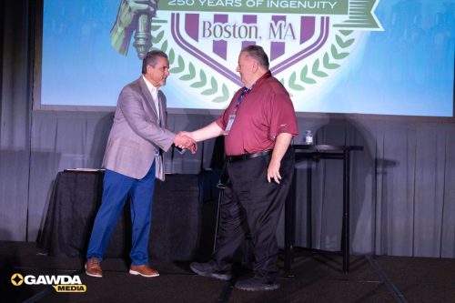 Two men shake hands on a stage during an award or recognition moment, with a Boston, MA crest on a screen behind them and GAWDA Media logo visible in the corner.
