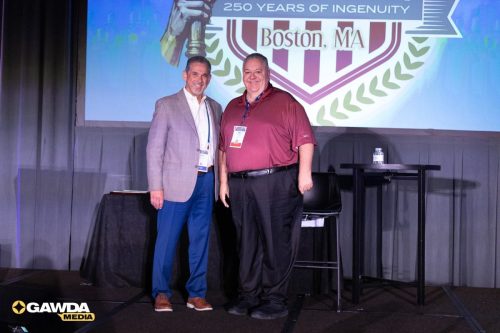 Two men pose on a conference stage, smiling, with badges around their necks; a large crest and banner are projected behind them.