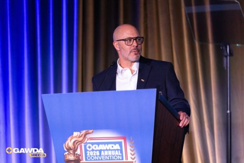 Bald man with glasses in a navy suit speaking at a podium on a conference stage (GAWDA 2026 convention banner visible).