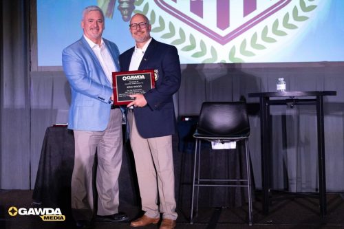Two men in suits on a stage exchange a plaque during an awards ceremony, with a large banner behind and the GAWDA Media logo in the bottom corner.”,